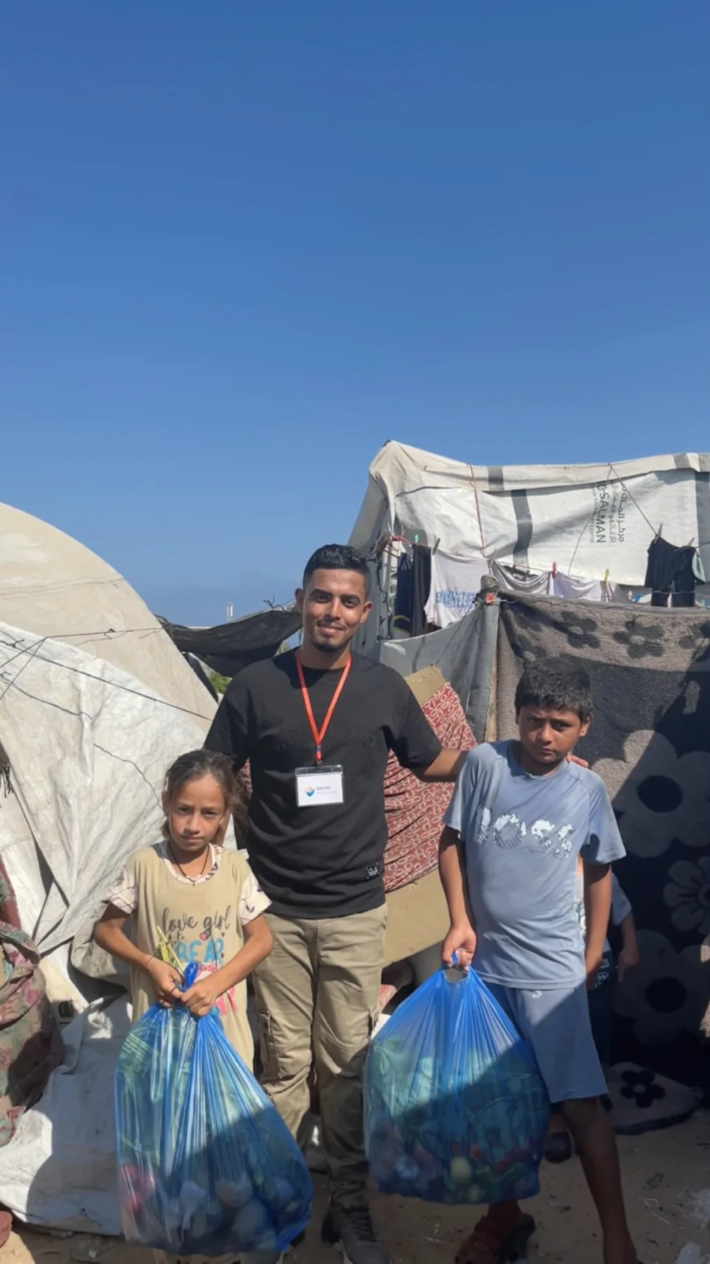 Two children holding food assistance bags beside tents